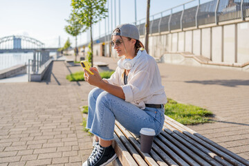 Young Hispanic hipster girl browsing mobile phone while sitting in street with takeaway beverage. 