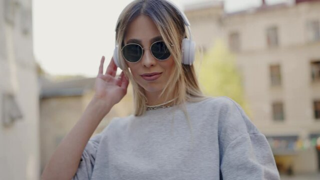 Young woman in stylish sunglasses listens to music in headphones against old buildings. Blonde woman tourist enjoys music sound in fresh air during observing city. Music lover on vacation