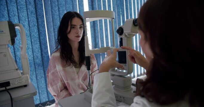 Young woman at female ophthalmologist appointment to get eyes checked in clinic. Oculist explains way of ophthalmometer operating to patient before procedure. Diagnosis of eye diseases