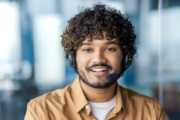 Portrait of positive indian guy wearing headset with microphone and looking at camera on blurred...