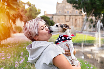 Woman with short grey hair holding and kissed on cheek by small senior dog in park outdoor. Dog is...