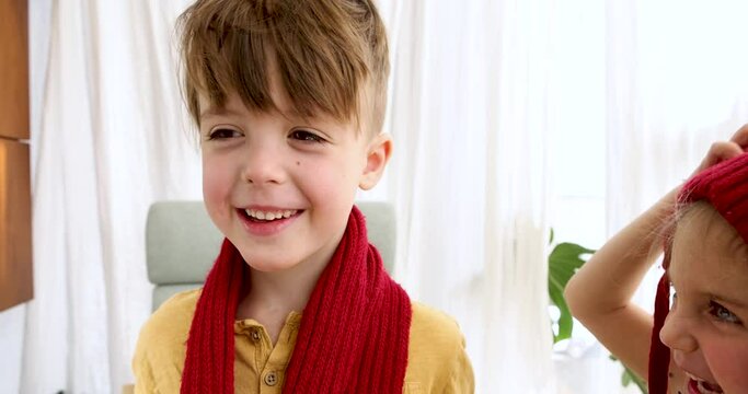 Little girl hanging a dark red scarf on her brother boy at home close-up portrait. Two cute little cheerful children are playing at home. Children Winter activities for children. Knitted scarf, hat