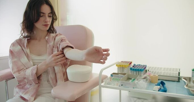 Woman sits in armchair in modern medical laboratory to collect blood for tests. Woman pulls shirt sleeve down over arm for injection site covered with band-aid. Diagnostic session
