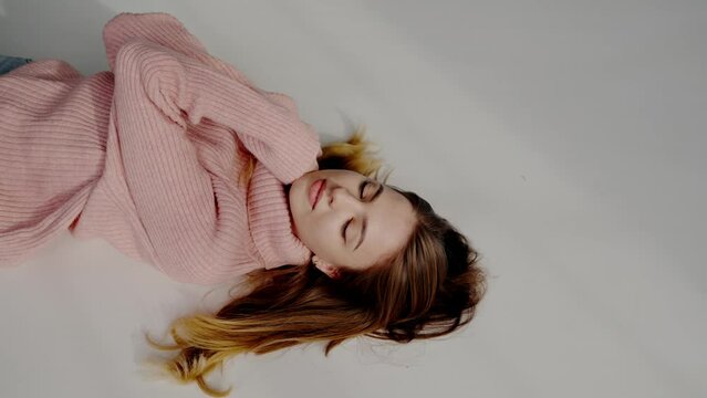 Young woman in sweater poses lying on gray floor with loose hair for photo. Young woman demonstrates professional skills during photo session for fashion magazine. Artistic photo-shoot