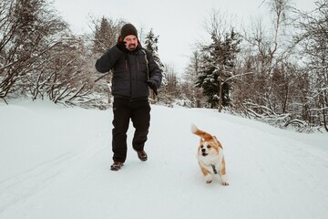 man and a corgi dog running through a winter snowy forest