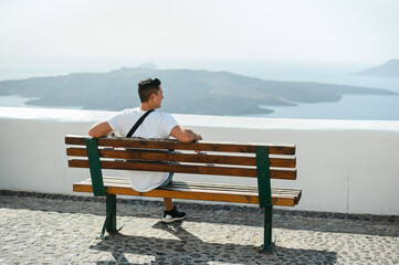 Back view of young man sitting on bench looking at Santorini coastline - obrazy, fototapety, plakaty