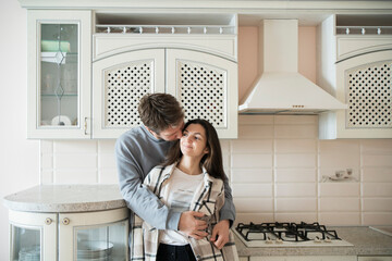 A couple stands close together in an affectionate embrace, exchanging loving looks in the comfort of their sunlit kitchen. - Powered by Adobe