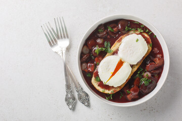 Oeufs en meurette is a classic French dish of poached eggs covered in a rich red wine sauce filled with lardons, mushrooms and onion closeup on the plate on the table. Horizontal top view from above