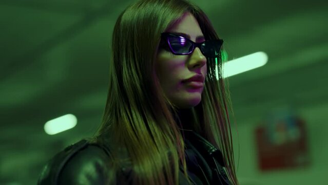 Young woman model with long loose hair in sunglasses poses in underground parking. Woman in leather jacket creates captivating visual juxtaposition against parked cars. Stylish girl