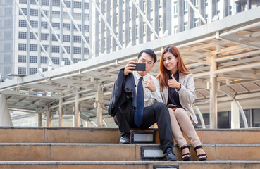 A joyful businessman and woman taking a selfie while sitting together on the stairs, Young Asian business people team discussing in the morning