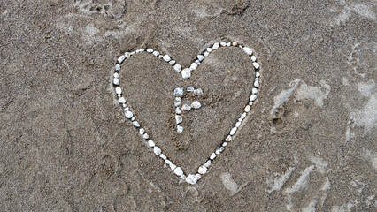 Beautiful, romantic and artistic love heart shape made with white pebbles on beach and the letter F inside representing Love Forever