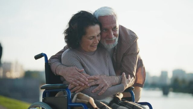 Husband walking with wife in wheelchair. Senior family couple in city park near pond. Elderly man hugging support woman showing love care, they talk smiling. Living with physical disability concept.