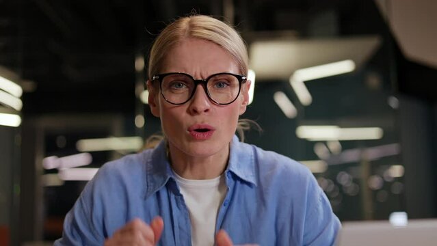 Blond woman putting on glasses and looking at computer screen with surprised look while sitting in coworking space. Glad female dressed in blue shirt saying wow and staring with amazement at camera.