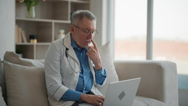 Professional serious senior doctor works on laptop sits in his modern office in hospital. Therapist doing paperwork, working with electronic documents, patients cards. Job old age practice concept.