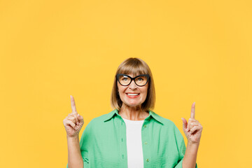 Elderly blonde woman 50s year old wear green shirt glasses casual clothes point index finger overhead on area indicate on area copy space mock up isolated on plain yellow background Lifestyle concept - obrazy, fototapety, plakaty