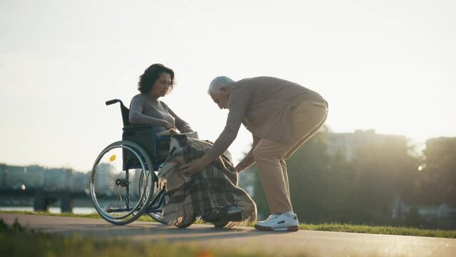 Husband walking with wife in wheelchair. Senior family couple spending time in city park near pond. Man helped woman showing love care, they talking. Living with physical disability concept.