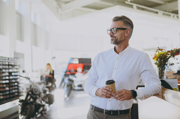 Male successful businessman man wear classic shirt prop up chin look aside drink coffee stand at office table in dealership store indoors want to buy new motorbike or car. Business lifestyle concept..