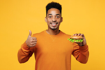 Young smiling man wear orange sweatshirt casual clothes hold eat burger show thumb up gesture...