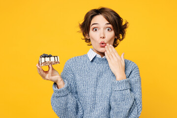 Young shocked surprised woman wears grey knitted sweater shirt casual clothes hold in hand eat piece of sweet cake cover mouth with hand isolated on plain yellow background studio. Lifestyle concept.