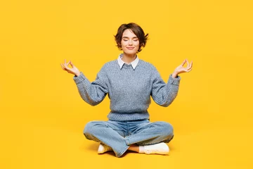  Full body young woman wears grey knitted sweater casual clothes sits hold spreading hands in yoga om aum gesture relax meditate try to calm down isolated on plain yellow background. Lifestyle concept. © ViDi Studio