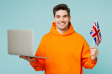 Young smiling IT man he wears orange hoody casual clothes hold British flag use work on laptop pc computer isolated on plain pastel light blue cyan color background studio portrait. Lifestyle concept. - obrazy, fototapety, plakaty