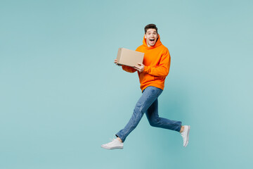 Full body surprised excited young man he wearing orange hoody casual clothes hold cardboard box jump high isolated on plain pastel light blue cyan color background studio portrait. Lifestyle concept.