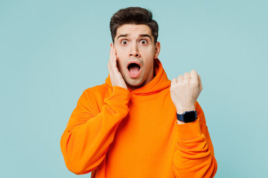 Young shocked scared sad astonished man he wearing orange hoody casual clothes showing time on smart watch isolated on plain pastel light blue cyan color background studio portrait. Lifestyle concept. - Powered by Adobe