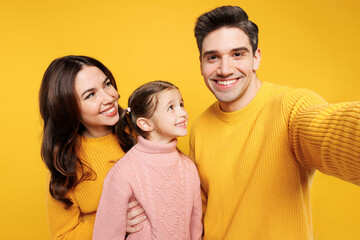 Close up young fun happy parents mom dad with child kid girl 7-8 years old wear pink sweater casual clothes do selfie shot on mobile cell phone isolated on plain yellow background. Family day concept.