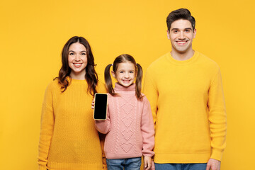 Young smiling parents mom dad with child kid girl 7-8 years old wear pink sweater casual clothes hold use blank screen area mobile cell phone isolated on plain yellow background. Family day concept.
