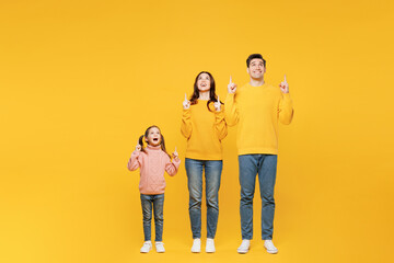 Full body young happy parents mom dad with child kid girl 7-8 years old wear pink sweater casual clothes point index finger overhead on area isolated on plain yellow background. Family day concept. - obrazy, fototapety, plakaty