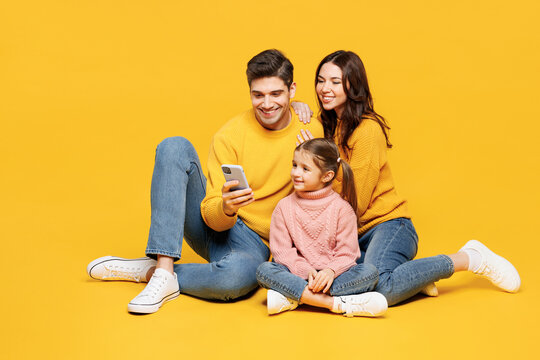 Full body young smiling happy parents mom dad with child kid girl 7-8 years old wear pink sweater casual clothes sitting use mobile cell phone isolated on plain yellow background. Family day concept. - Powered by Adobe
