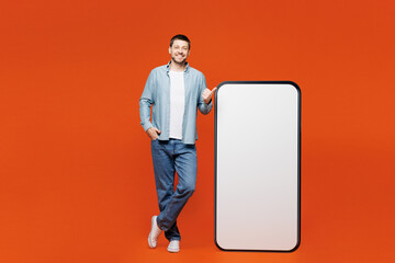 Full body young man wears blue shirt white t-shirt casual clothes point thumb finger on big huge blank screen mobile cell phone smartphone with workspace area isolated on plain red orange background.