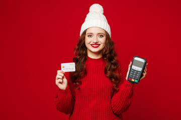 Young woman wears knitted sweater white hat casual clothes hold wireless modern bank payment terminal to process acquire credit card isolated on plain red color background studio. Lifestyle concept.