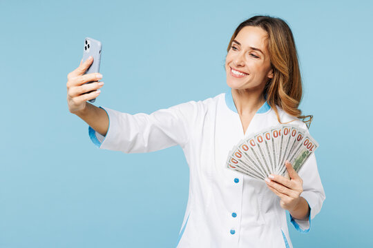 Doctor woman wear white medical gown suit work in hospital clinic office hold fan cash money dollars do selfie shot on mobile cell phone isolated on plain blue background Health care medicine concept