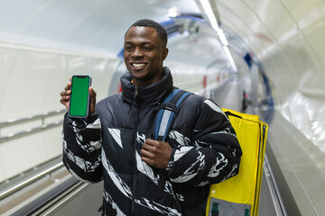 An African-American courier with a yellow backpack outside with a mobile phone with a green screen