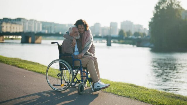 Wife walking with husband in wheelchair. Senior family couple spending time in city park near river. Woman hugging man showing love care, they smiling. Living with physical disability concept.