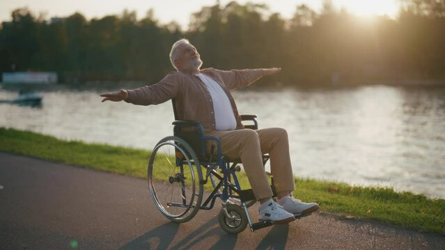 Senior happy man in wheelchair enjoys freedom. Paralysis male with special need spreading arms on sides breathing fresh air walking in city park. Living with physical disability in old age concept.