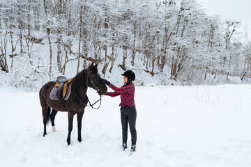 A beautiful brunette girl in a plaid black-and-white shirt walks with a big black horse in a snowy...