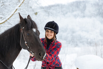 A beautiful brunette girl in a plaid black-and-white shirt walks with a big black horse in a snowy...