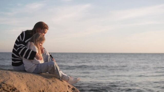 Calm mother with little daughter sitting on a rock by the sea in the evening, Spain, Alicante