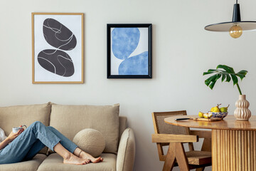 Warm and cozy interior of living room space with two mock up poster frames, round table, leaf in vase, chair, pedant lamp, decoration and woman lying on the sofa. Minimalist home decor. Template. - obrazy, fototapety, plakaty
