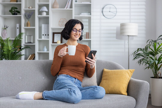 Smiling young woman sitting on sofa at home and resting, using mobile phone and holding cup with drink