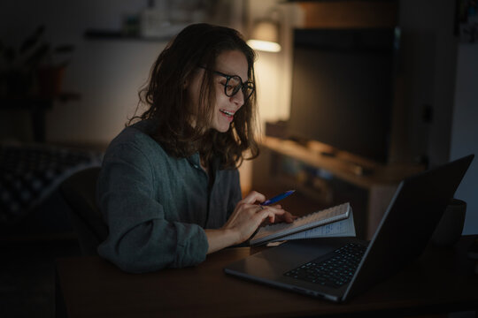 Young woman with notes in front of laptop at home in the evening, online courses