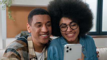 Happy African American smiling couple man woman at home carefree talking at mobile phone webcam...