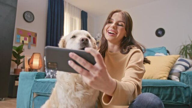 Positive Caucasian woman taking photo for new post in social media with her cute doggy. Pretty female holding smartphone and joyfully looking straight on camera while sitting on floor.