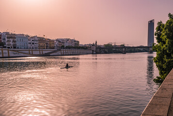 Guadalquivir river with silhouette of a canoeist, architecture of city, Isabel II bridge and Seville tower in the background at evening, SPAIN