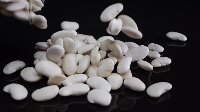 Gently falling white beans from glass containers onto a dark reflective base, showcasing their role in nutritious vegan meals.