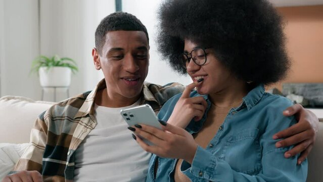 Happy married African American couple man and woman at home couch looking mobile phone shopping website app store internet using smartphone search booking tickets choosing purchase discussing talking