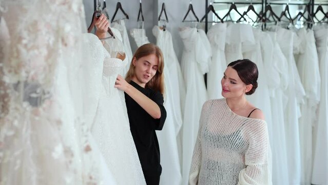 Young attractive girl choosing white dress with help of shop assistant at wedding salon.