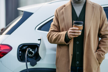 Close up of businessman waiting while electric car is charging, leaning against vehicle, drinking...
